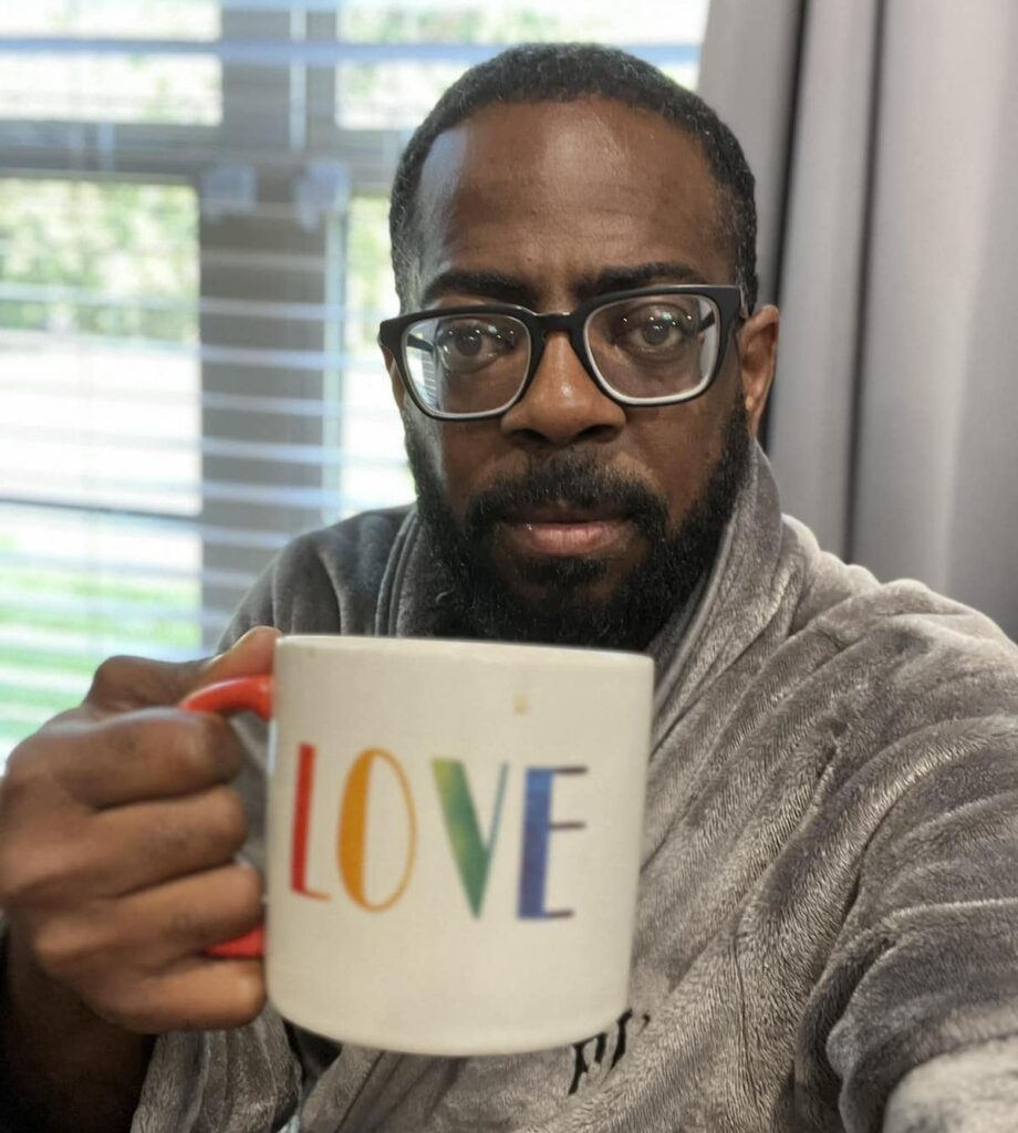 a photo of Fabian Nelson in a gray house robe sitting in front of a window holding a mug that says "LOVE" in rainbow colors