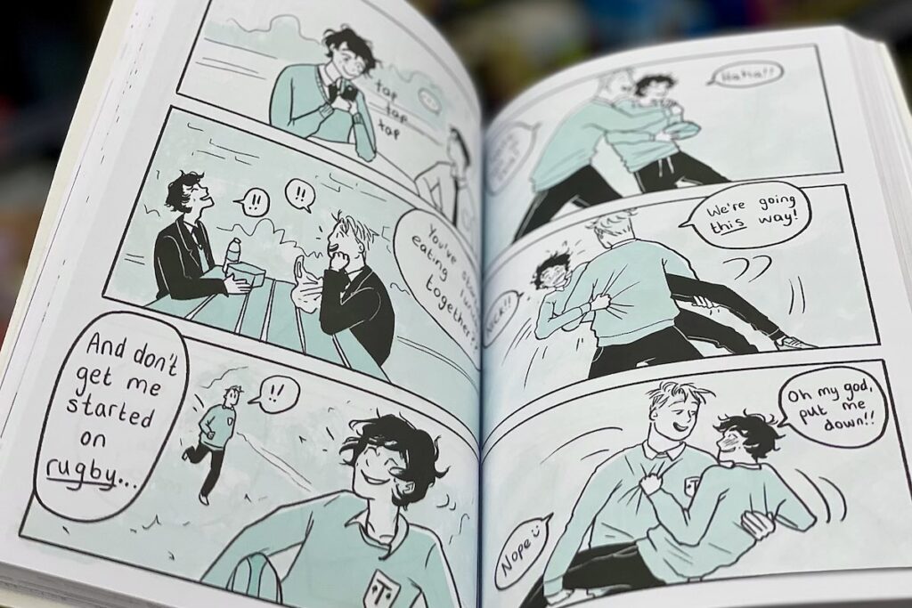photo shows twp pages of a graphic novel: In the first frame, a boy is sitting at a table typing on his phone and it says "tap tap tap"; in the second one, another boy sitting across from him as they both laugh says "You've been eating lunch together?" the third shows the boy from the first one walking and another running behind him that says "And don't get me started on rugby"; the fourth shows one of the boys tackling other as the two laugh; the fifth shows the boy that was running up from behind picking the other one up; the boy being picked up says "NICK!!" and Nick replies: "We're going this way!" In the sixth frame, Nick is holding Charlie in his arms and Charlie says "Oh my God, put me down!!"; Nick says "Nope :)"