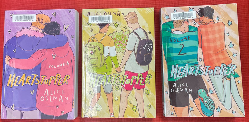 Photo shows three books: One says "Heartstopper Volume 4" by Alice Oseman and shows the backs of two boys with their arms around one another, heads close; the second shows the two boys from behind holding hands and wearing backpacks and says "Heartstopper Volume 3"; the third shows two boys lying down on their stomachs from above and looking at one another and says "Heartstopper volume 2"