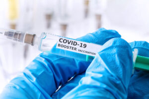 A blue gloved hand holds a syringe labeled "COVID-19 BOOSTER VACCINATION"