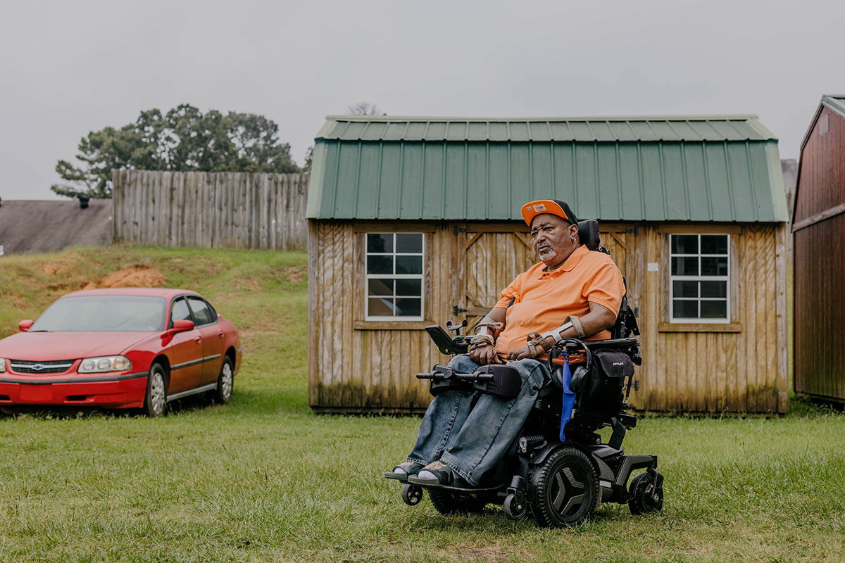 A full body of Dennis Mitchell, seated in a wheel chair, a shed visible behind him