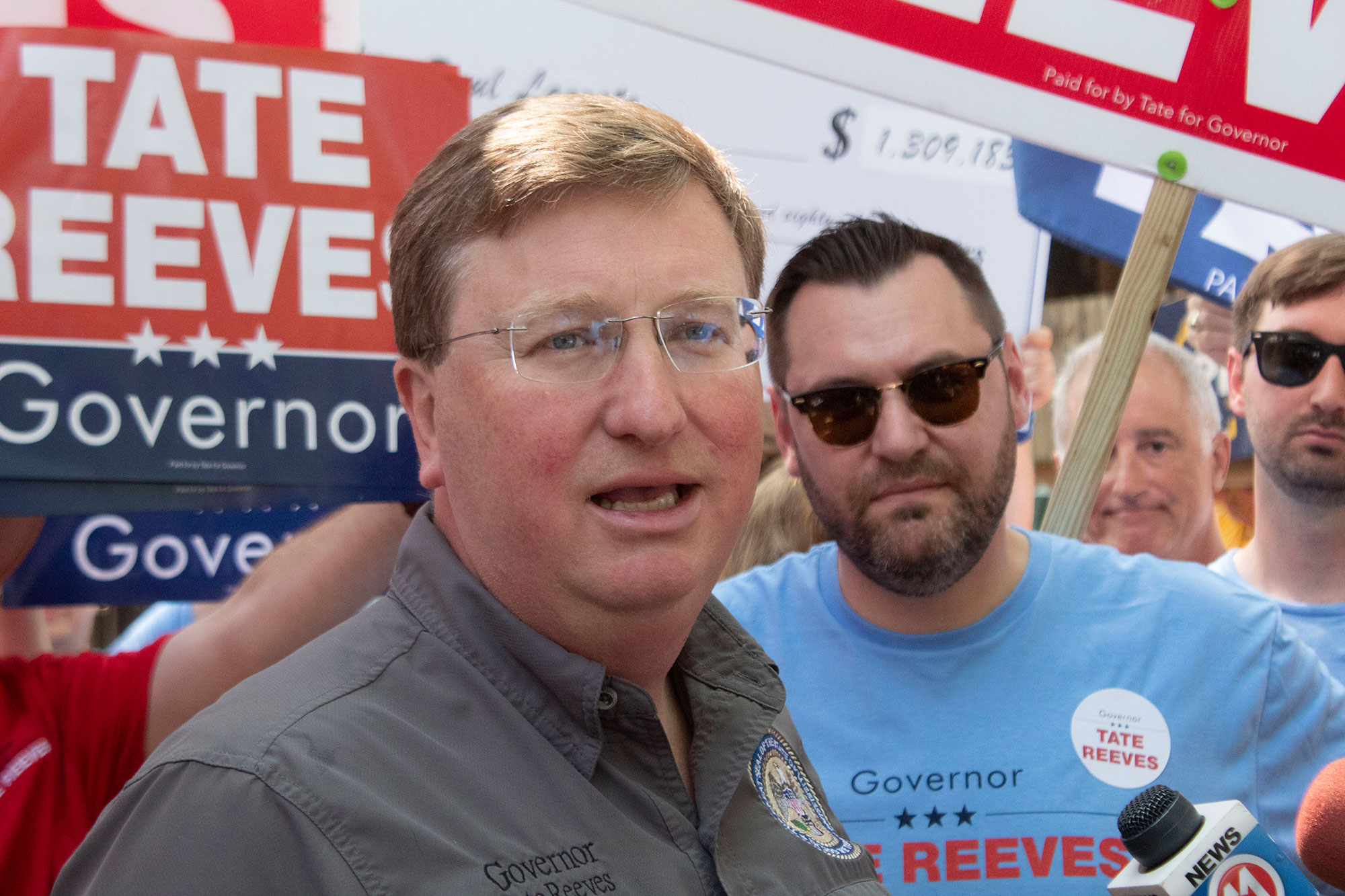 Tate Reeves speaking at a rally, as people hold signs with his name on it behind him