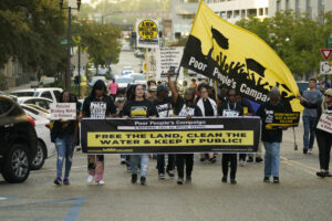 People parade down a street with a sign that reads "Free the Land, Clean the Water & Keep it Public!"