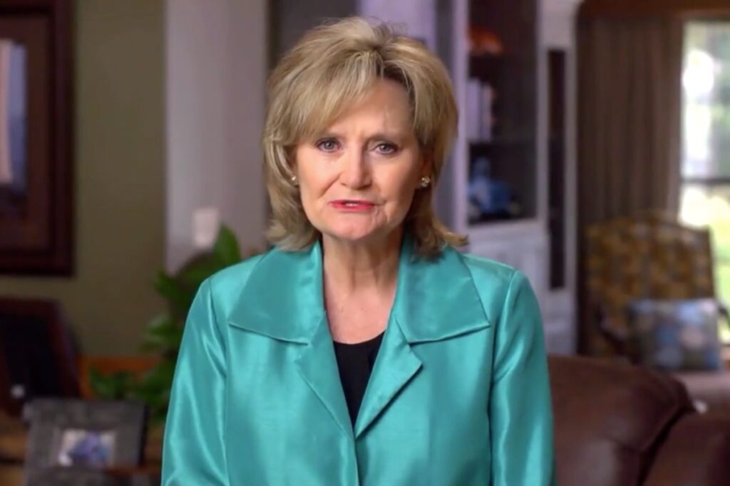 A screenshot of Cindy Hyde-Smith in a teal jacket speaking from inside an office