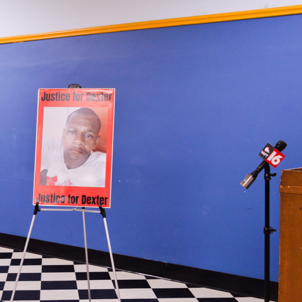 a photo of a poster standing next to a podium and WAPT microphone on a stand says, "JUSTICE FOR DEXTER" at the top and bottom with a photo of justice wade; the poster sits on an easel