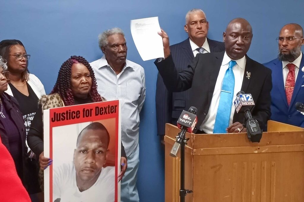 a photo of Ben Crump holding up a piece of paper while standing at a podium with half a dozen others standing behind and around him; to his left is Bettersten Wade, holding a poster with a photo of her son Dexter Wade's face with the words "JUSTICE FOR DEXTER" above his image