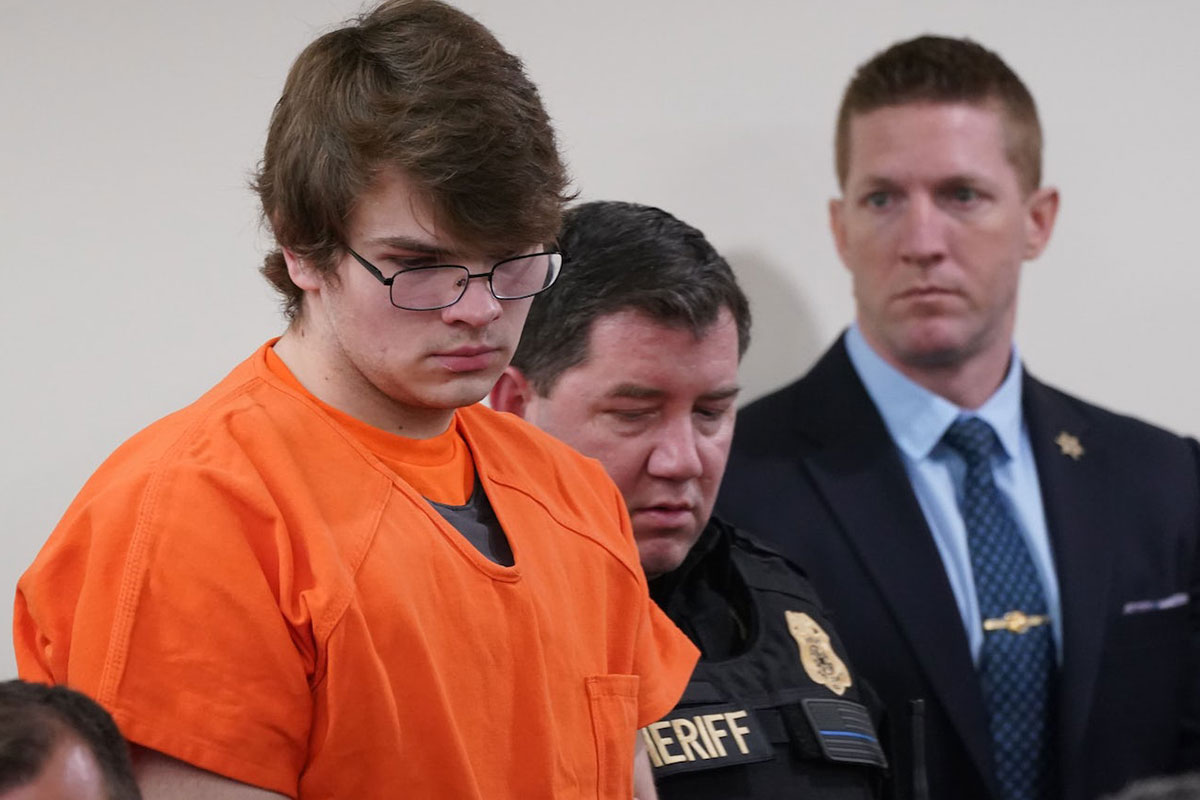 A man in orange jumpsuit and wrists restrained looks down at a court hearing