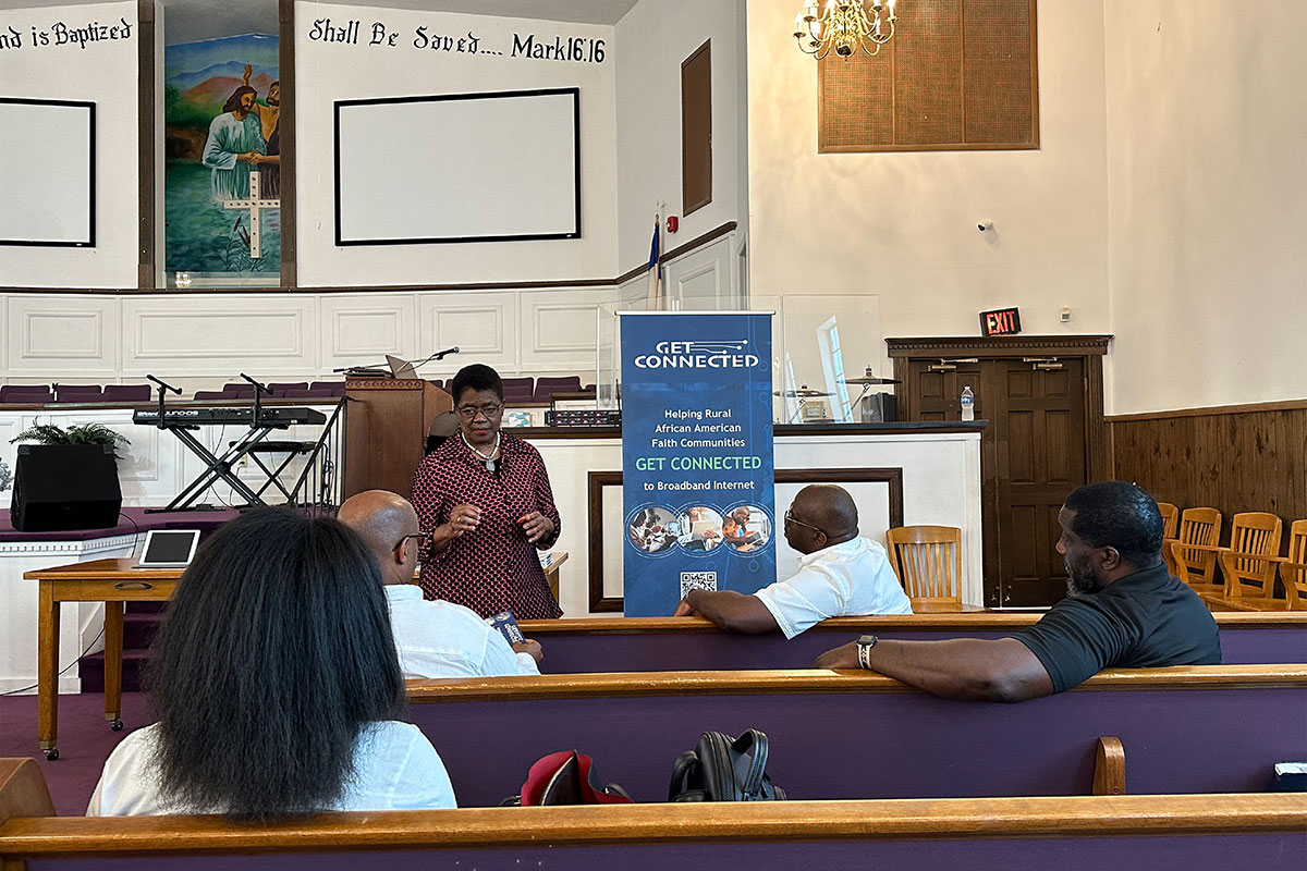 A woman speaks to people sitting in church pews. A sign beside her says "Get connected"
