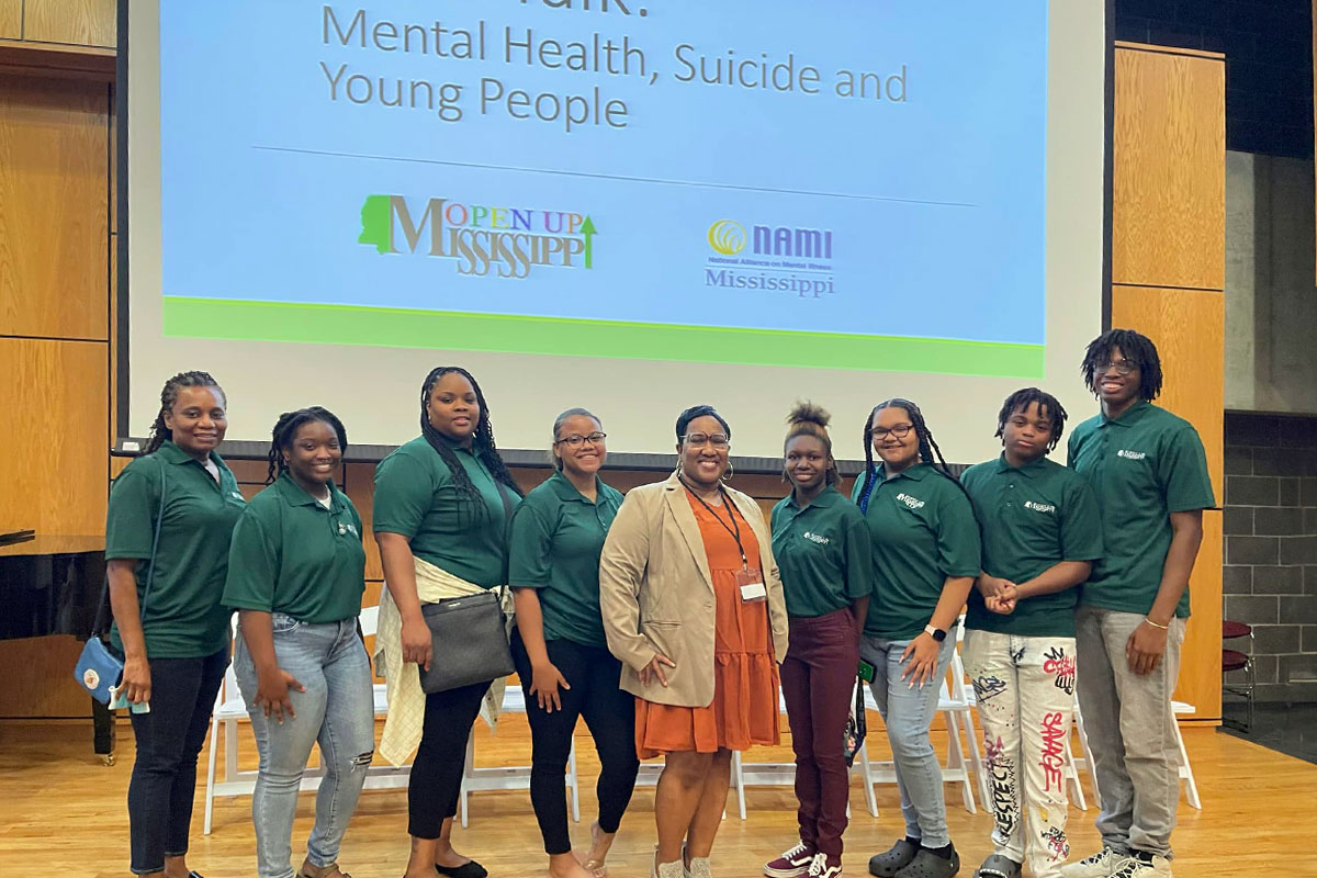 Shalonda Carlisle stands on state with several people in green shirts. A screen behind them reads "Mental Health, Suicide and Young People"