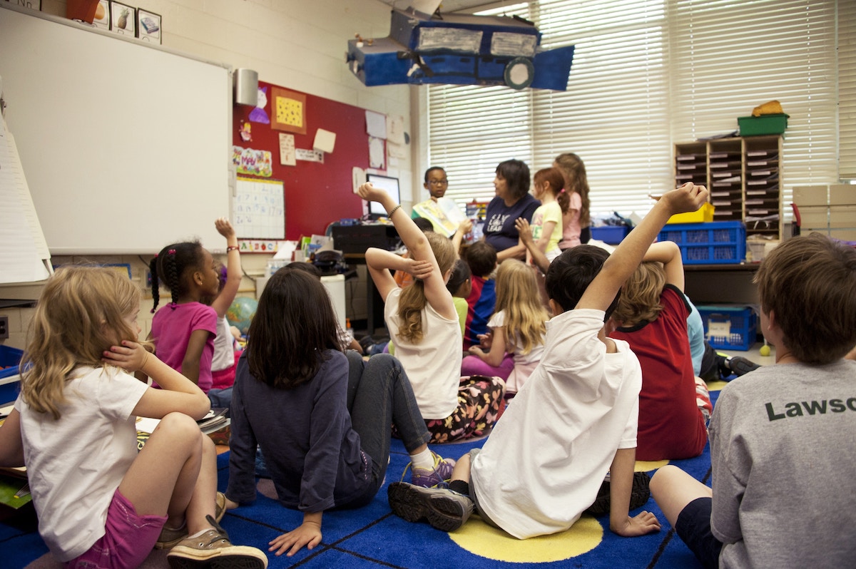 Several young students sit on the floor and raise their hands for the teacher