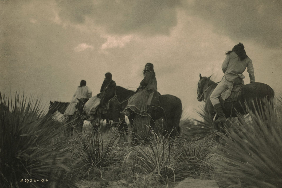 A line of Apache Indians riding horses across desert scrub land (land acknowledgement)