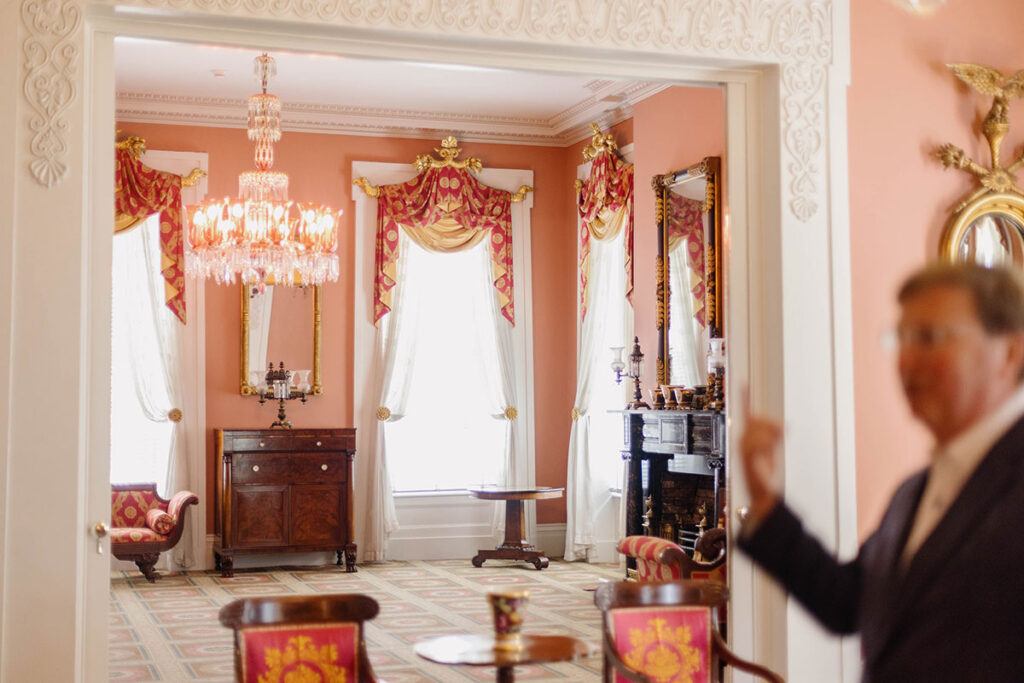 a photo shows Gov. Reeves pointing into a pink room with a chanedlier and large ornate windows and furniture