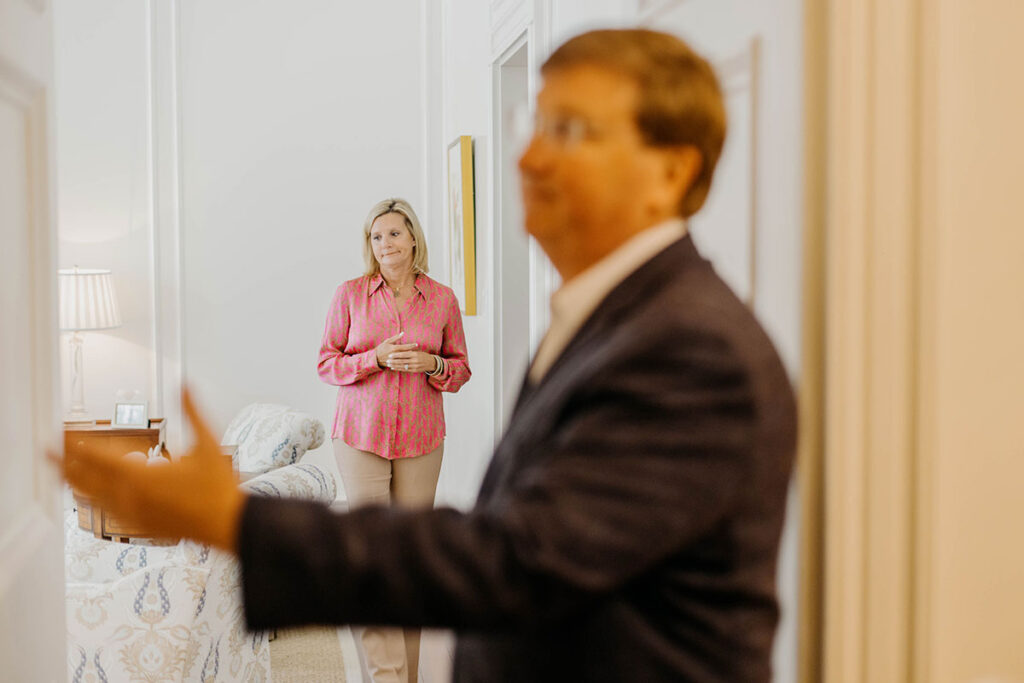 a photo of Gov. tate Reeves close up with his arm extended, Elee Reeves standing in the background looking towward him in a white room