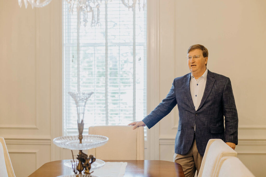 a photo of Tate Reeves standing at the end chair of a dining table in a large white room