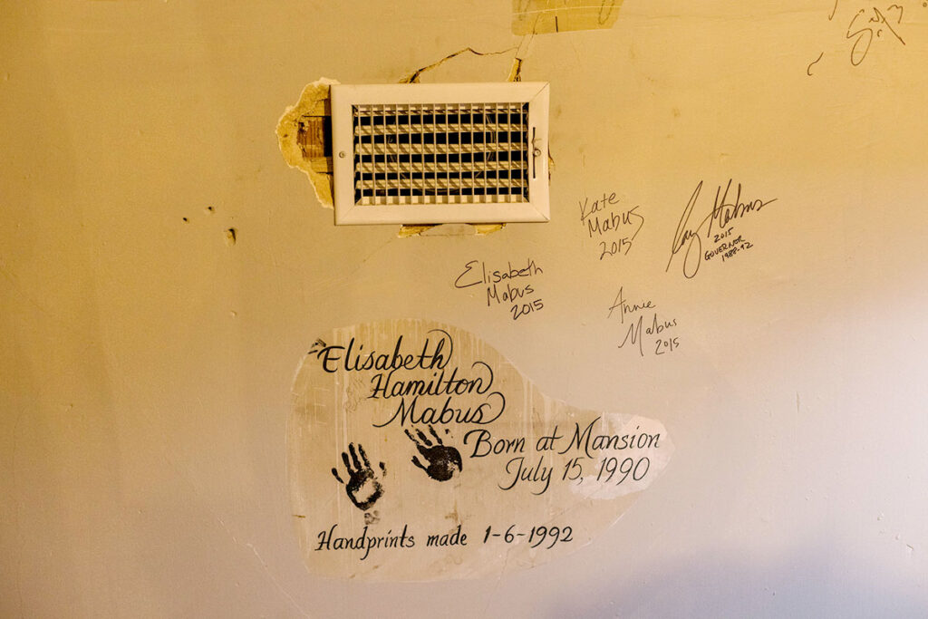Photo shows a dilapidated wall with an air vent with the wall crumbling around it. Signatures around the wall say: “Kate Mabus 2015” “Elizabeth Mabus 2015” “Ray Mabus - 2015 Governor 1988-92” “Annie Mabus 2015” “Elisabeth Hamilton Mabus - born at mansion July 15, 1990 - hand prints made 1-6-1992” includes a set of black ink handprints
