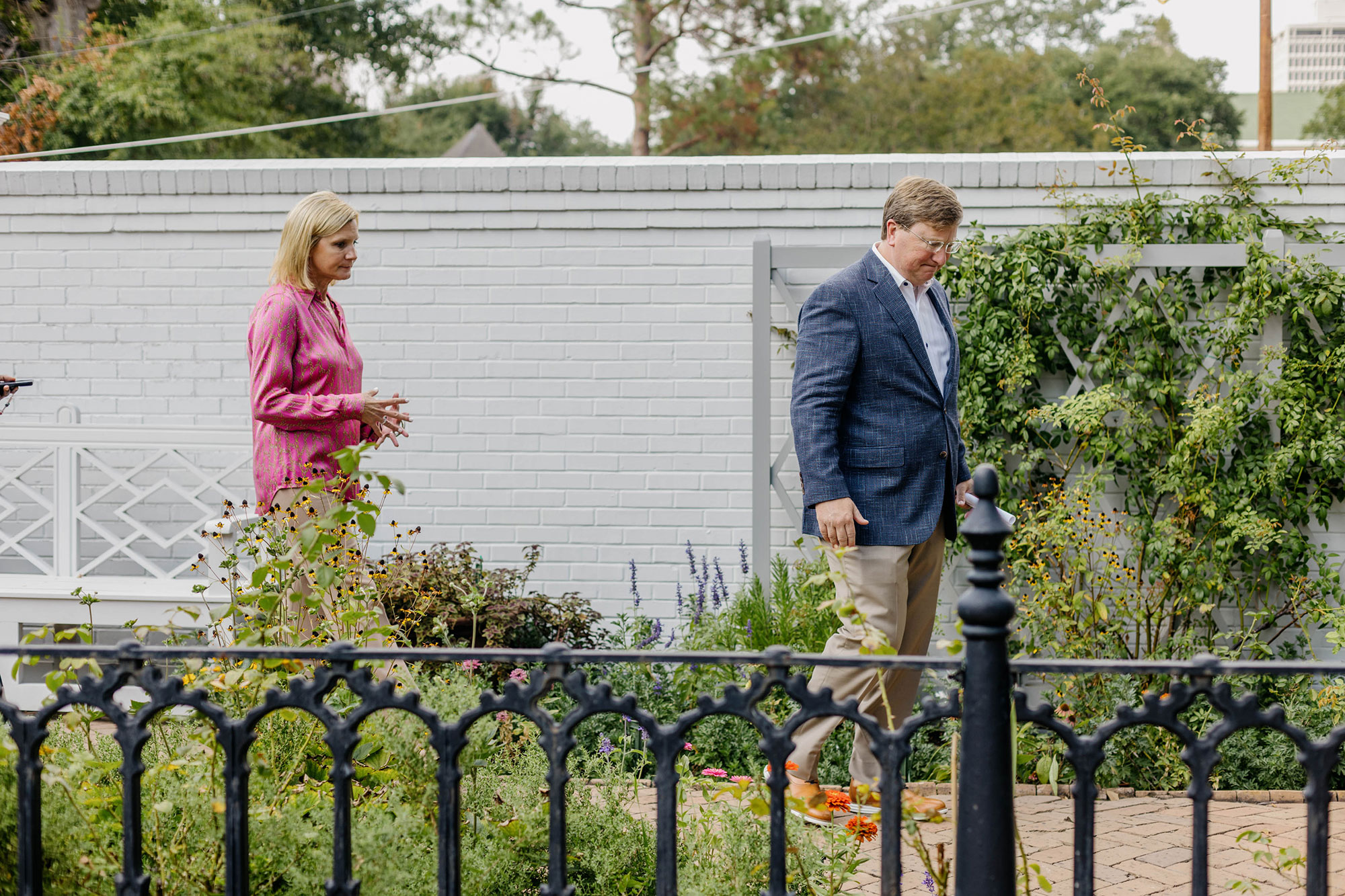 photo shows Tate Reeves and Elee Reeves walking through the garden of the governor's mansion, a white wall behind them