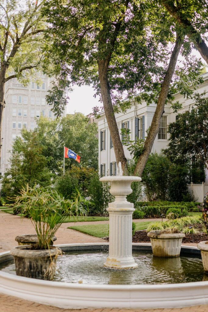 a photo of a fountain outside underneath trees with the governor's mansion in the background; a state flag flies in the distance
