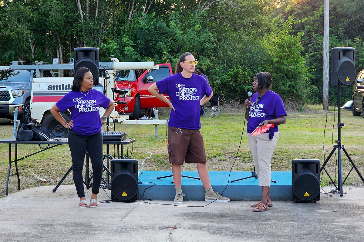 Three people in purple shirts that read One Nation One Project speaking on an outdoor stage