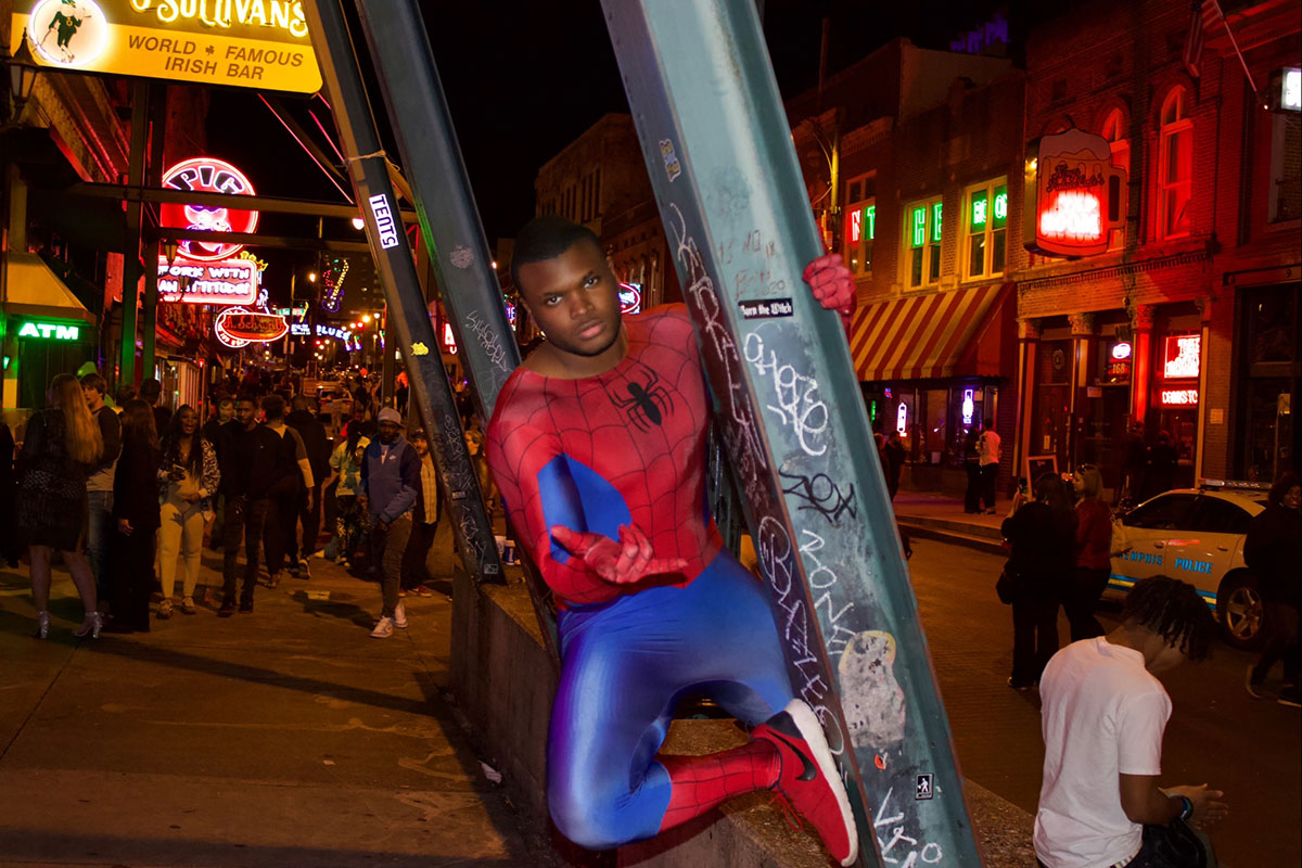 Preston Wallace dressed in the classic red and blue Spiderman costume, hanging from an iron girder on the sidewalk of a busy street