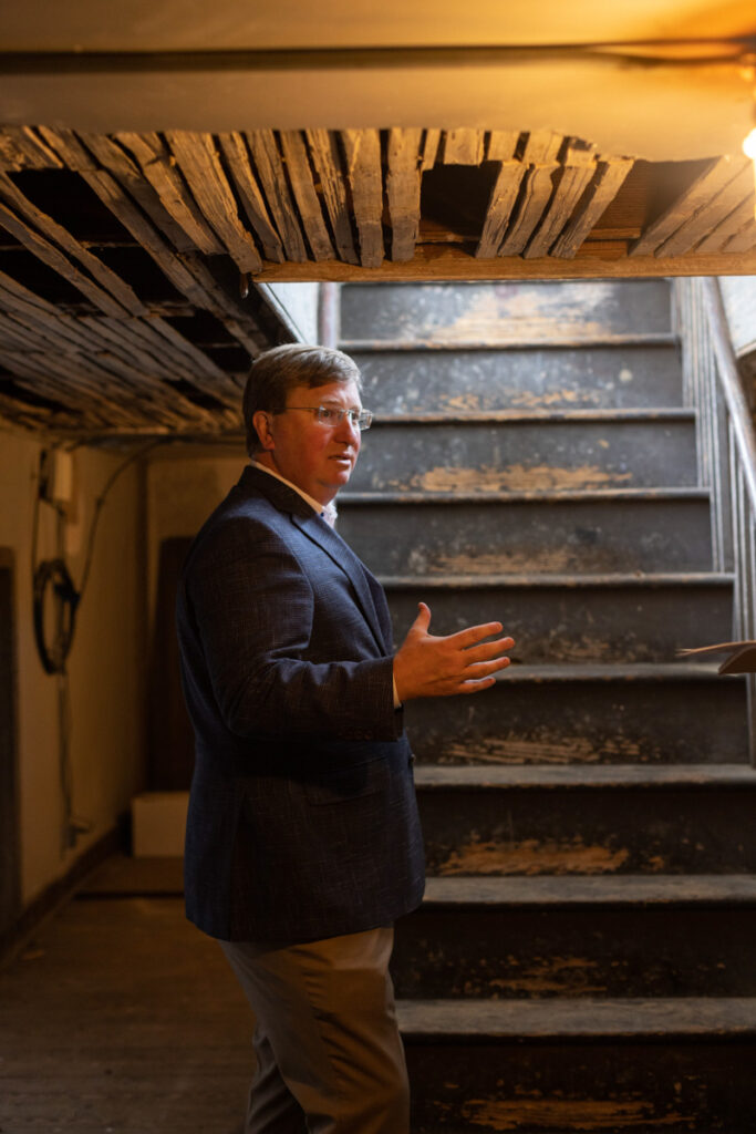 a photo of Tate Reeves standing near a rickety set of black stairs beneath exposed rickety wooden boards in the ceiling above him
