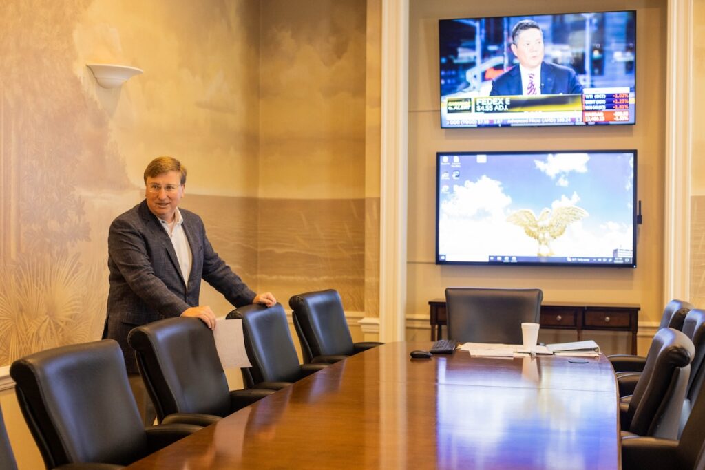 a photo of Tate Reeves standing at a conference desk, two large TVs on the wall behind him