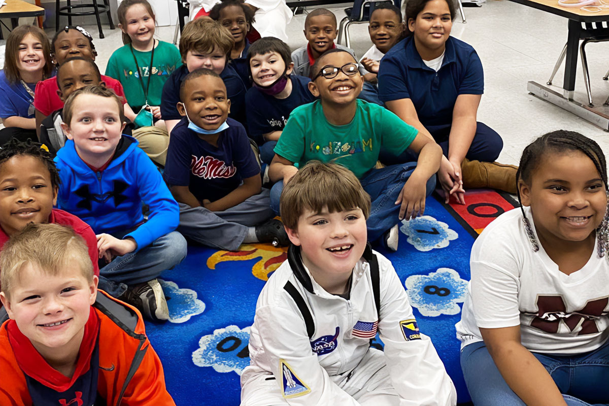Many children sit on a number mat on the floor, smiling upward (Mississippi Medicaid)