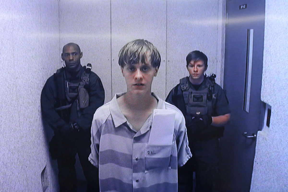 Video still of young blond man in prison jumpsuit surrounded by armed guards