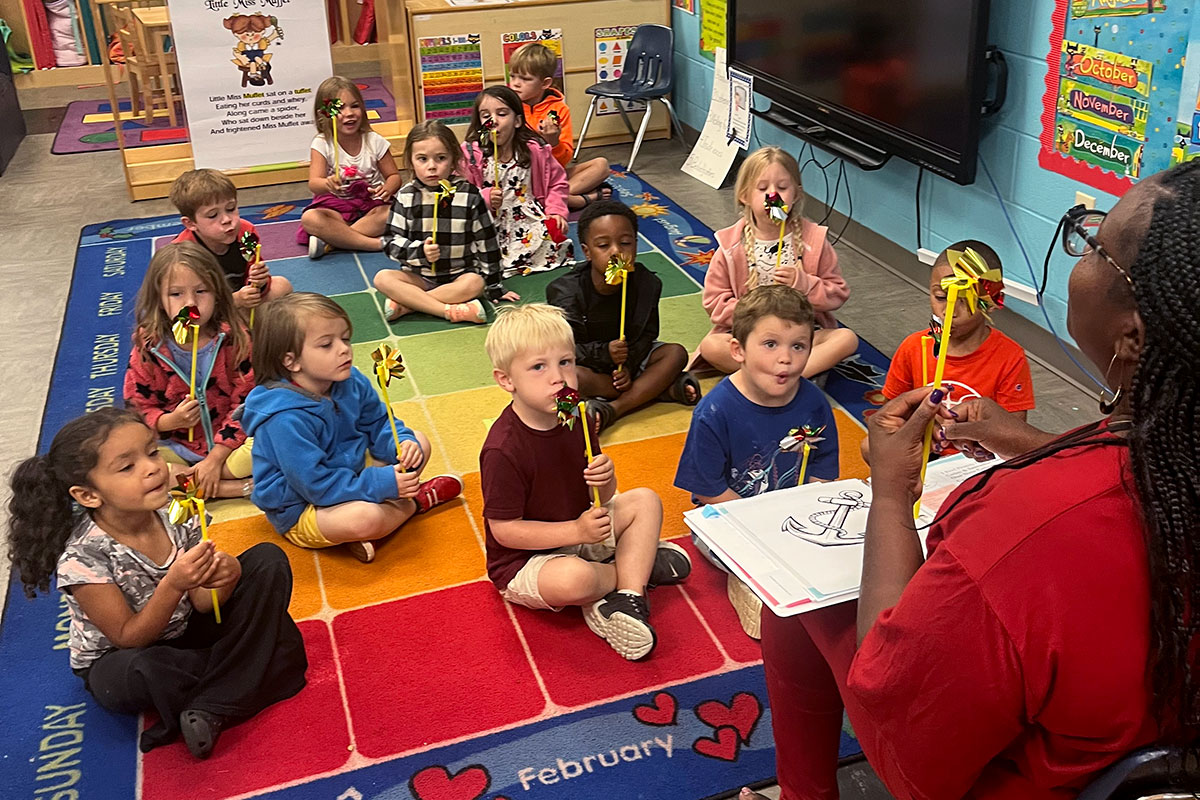 A teacher speaks to preschoolers sitting on colorful floor tiles