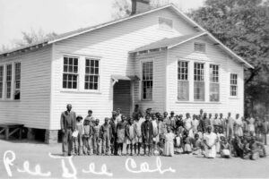 An old schoolhouse with a long row of Black adults and children posed for the photo in front