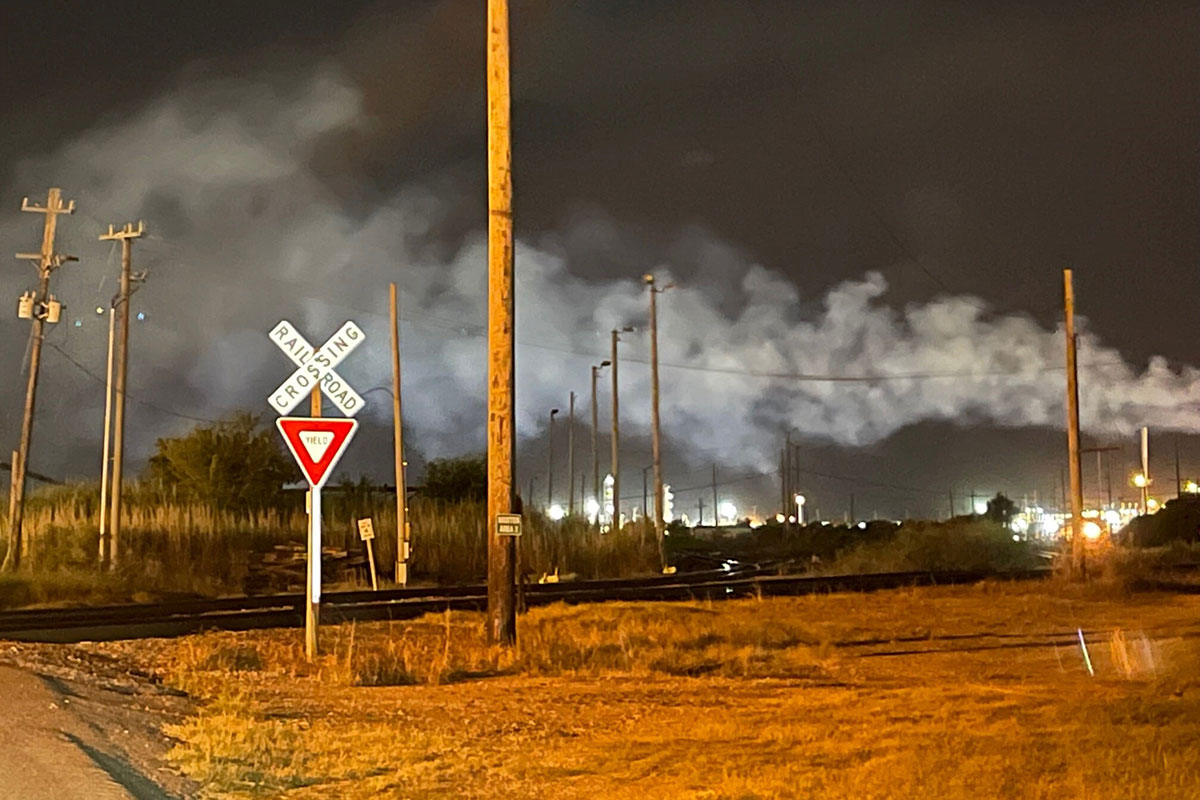 A rail road crossing at night with a Calcining plant visible in the background