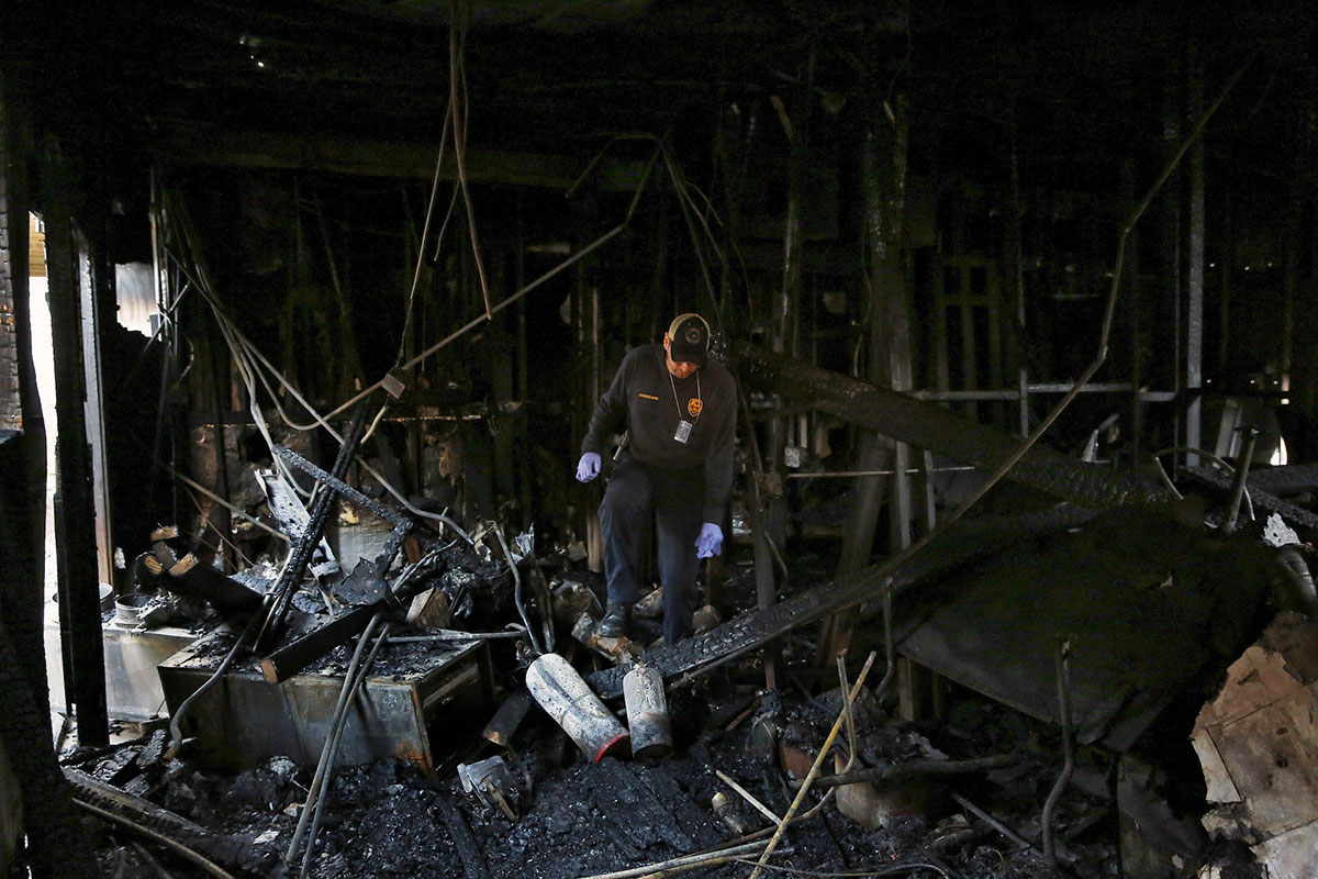 Fire inspector walks through ruins of a charred building, looking at the ground