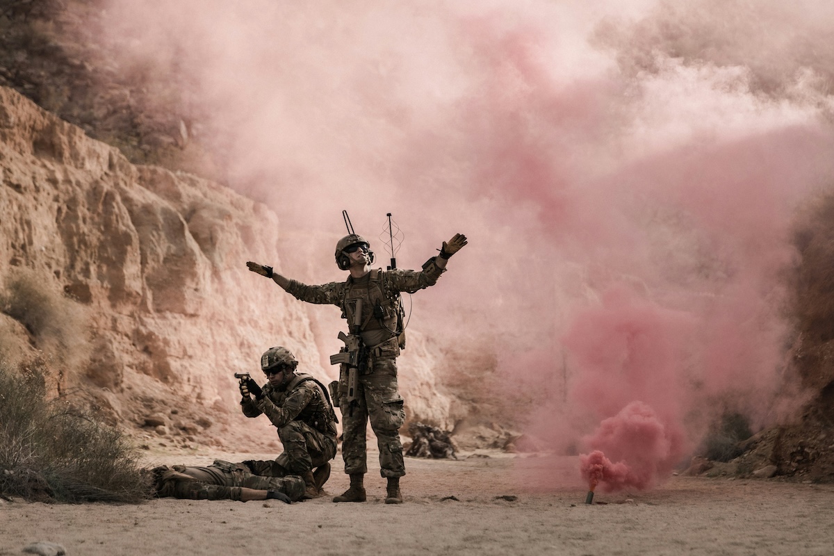 Two men in soldier gear stand in a sandy alcove with pink smoke behind them