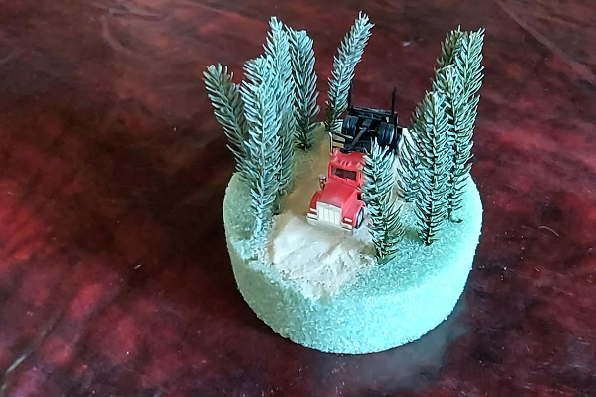 A small diaorama with a foam base shows a red truck diving down a white road, while tree stems stand is as trees to the sides