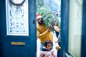 A man brings in a Christmas tree through front doors painted blue as a small girl walks ahead