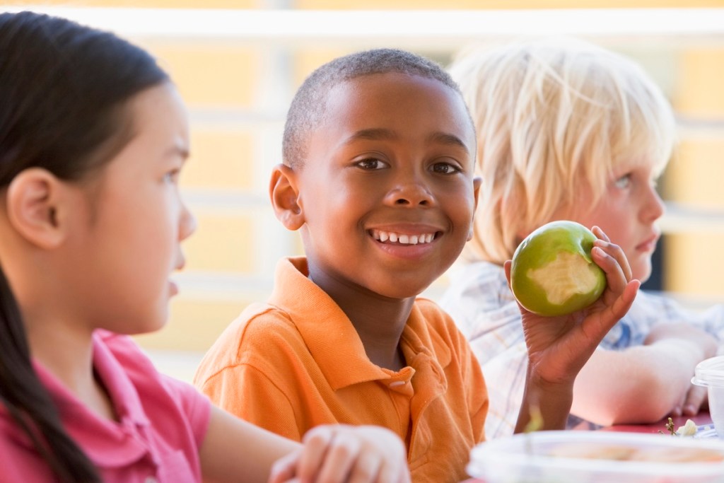 Kindergarten children eating lunch, one boy smiles while holding an apple