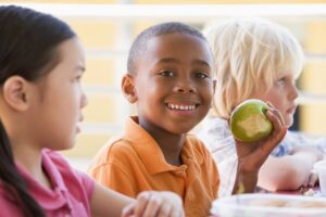 Kindergarten children eating lunch, one boy smiles while holding an apple