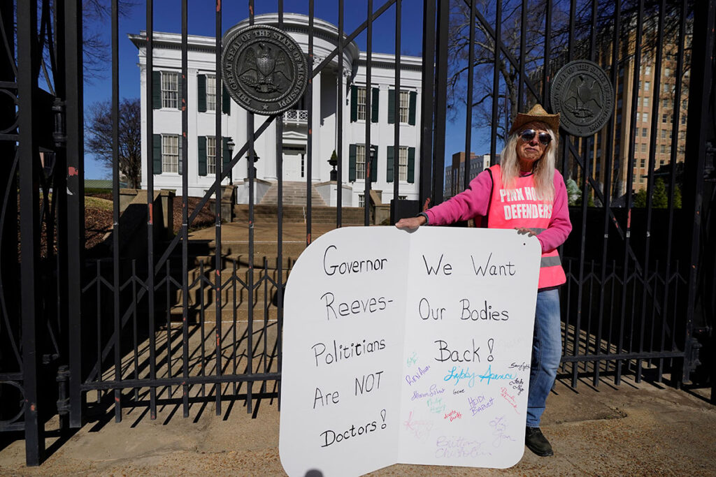 A woman in a pink shirt and vest that reads "Pink House Defenders" stands before the Mississippi Governor's Mansion