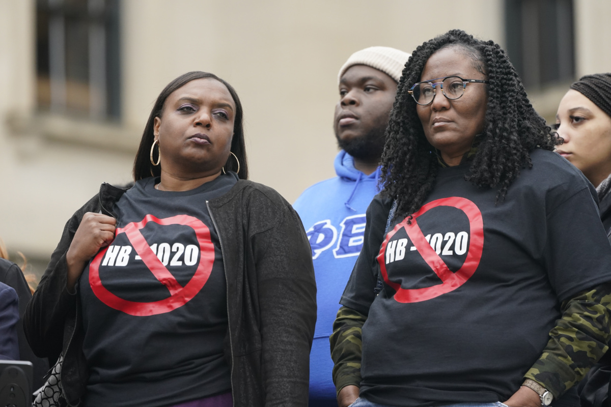 Two women stand side by side in a crowd. They both are wearing black tshirts with the words HB-1020 struck out