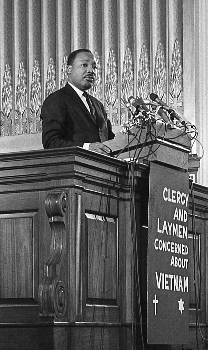 A Black man wearing a dark suit stands behind a lectern atop a sign that says clergy and laymen concerned about Vietnam