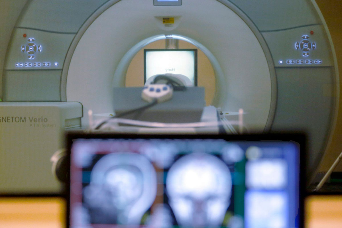 "A white MRI machine, which has a tube with a cot inside and a white monitor in the background, and a black monitor with blurred images of a human skull, in the foreground (helium)