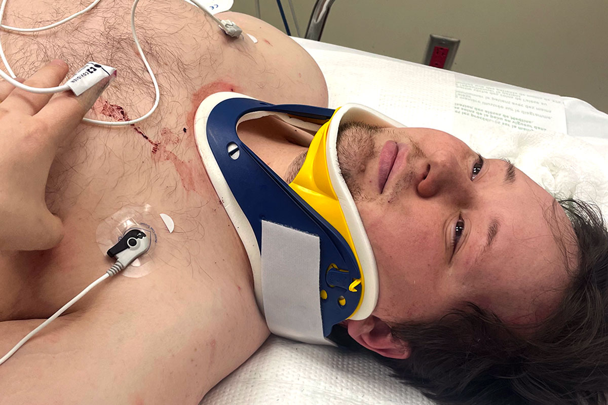 Nate laying in a hospital bed with a collar around his neck and wires attached to his chest and hand