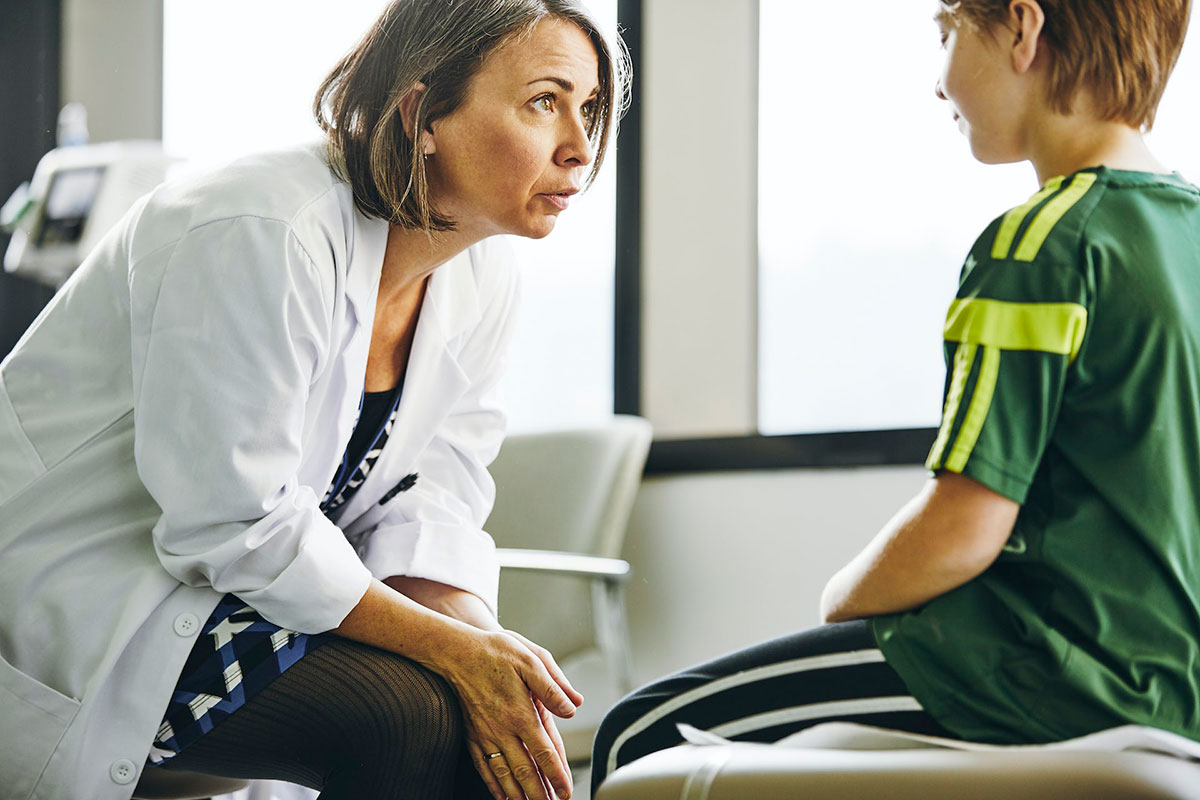 A woman in a white medical coat leans forward, seated, as she talks seriously with a seated boy in a green t-shirt. (patients)
