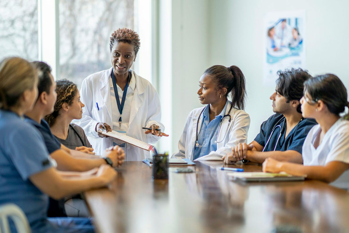 Six young adults sit at a conference table, some of them in scrubs, as a doctor in a white coat leads a discussion. (patients)