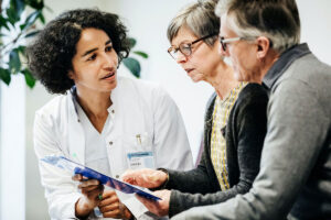 A doctor discusses something on a blue clipboard to two older patients