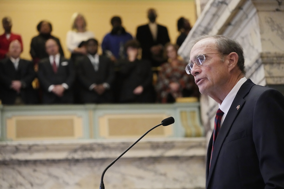 a photo of Delbert Hosemann shot from below of him standing at the microphone in the Senate chamber, people sitting in the galley above visible behind him above his head