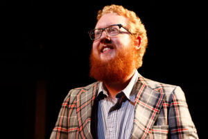 A bespectacled man with red hair and a beard wears a plaid jacket