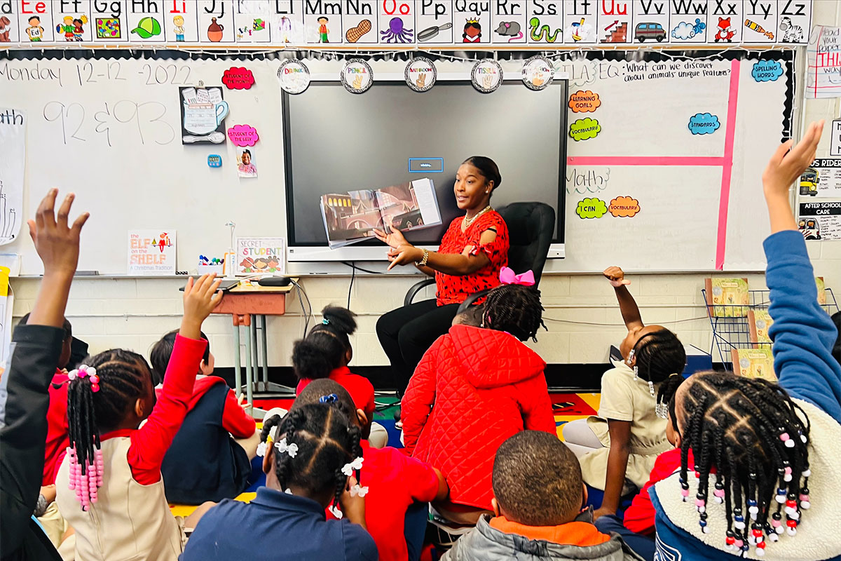 A teacher holds a book she's readying to a group of young children seated on the floor before her