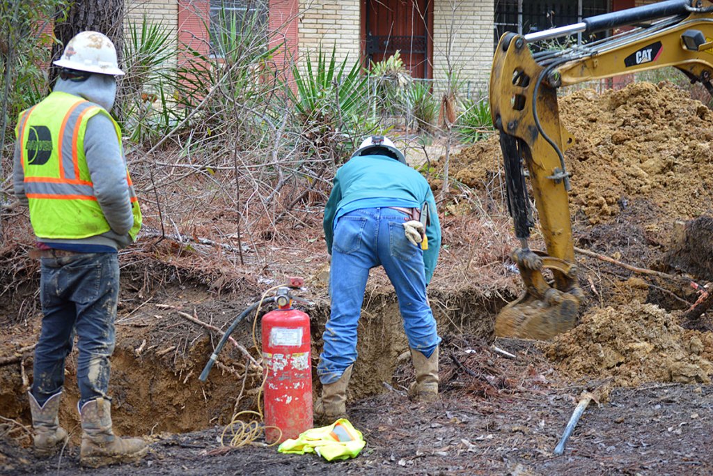 Two men in work gear look into a trench dug by a yellow digging device
