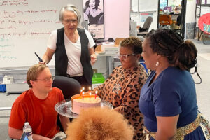 People stand around a seated man, presenting a birthday cake with lit candles