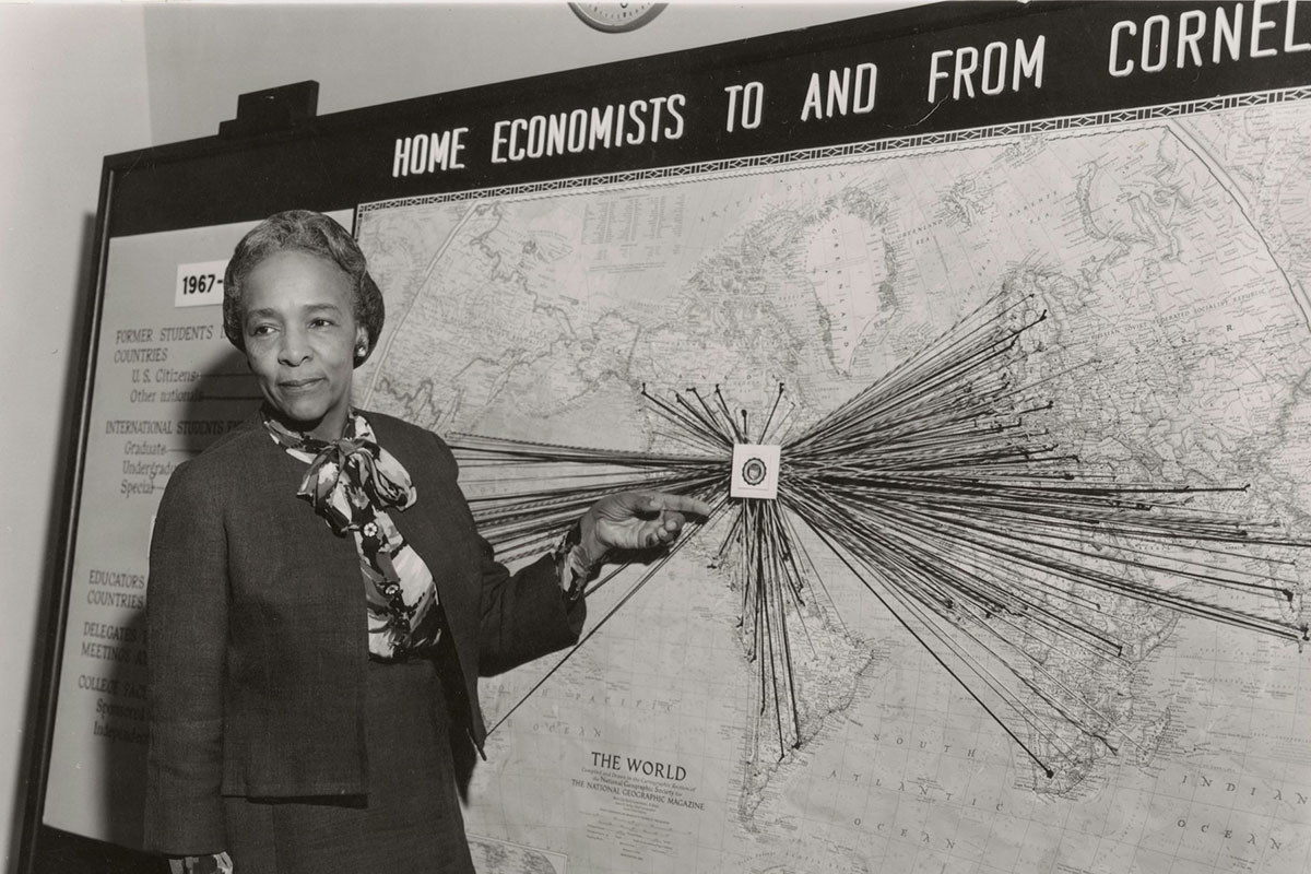A woman stands in front of a map of the world with pins radiating from Cornell University